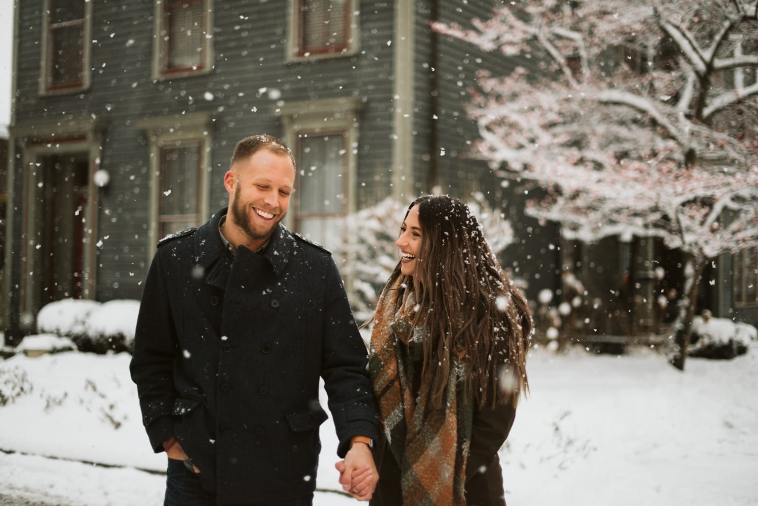 A couple laughing, walking through a historic neighborhood in Indianapolis as big snowflakes fall around them.