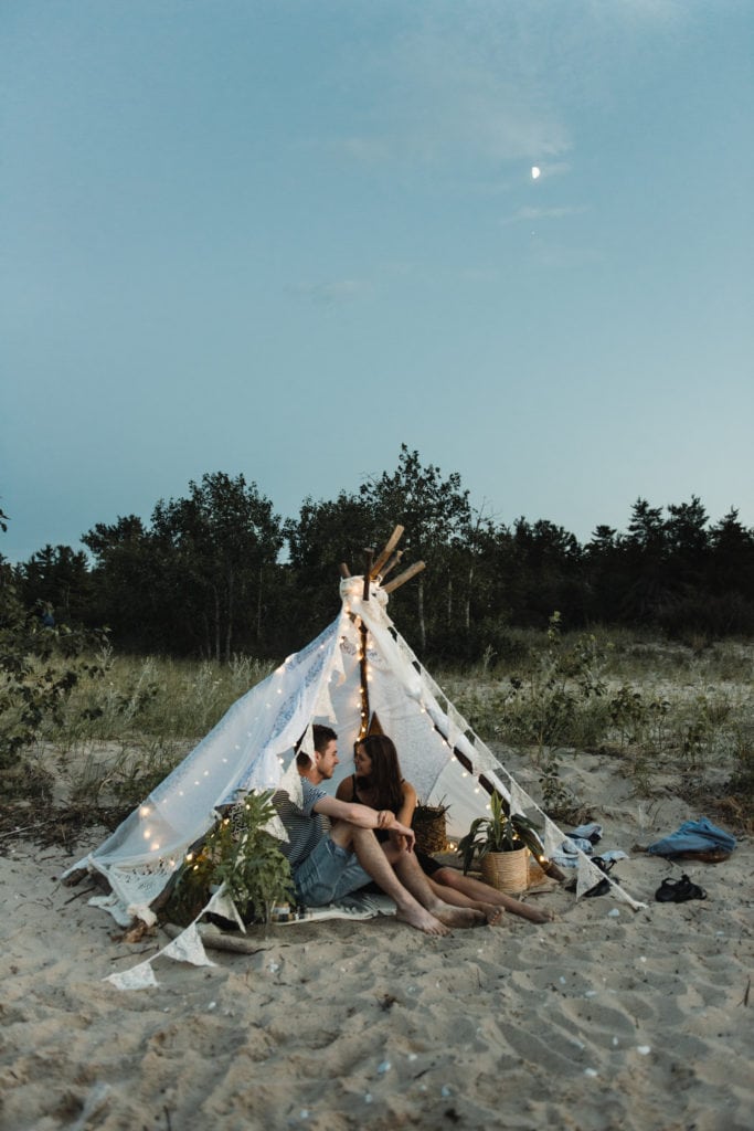 A couple smiling and embracing in a handmade teepee on the beach in Sleeping Bear Dunes, Michigan.