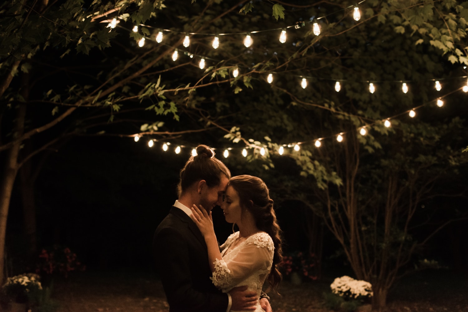 A bride and groom embrace underneath string lights after their intimate wedding in a forest.