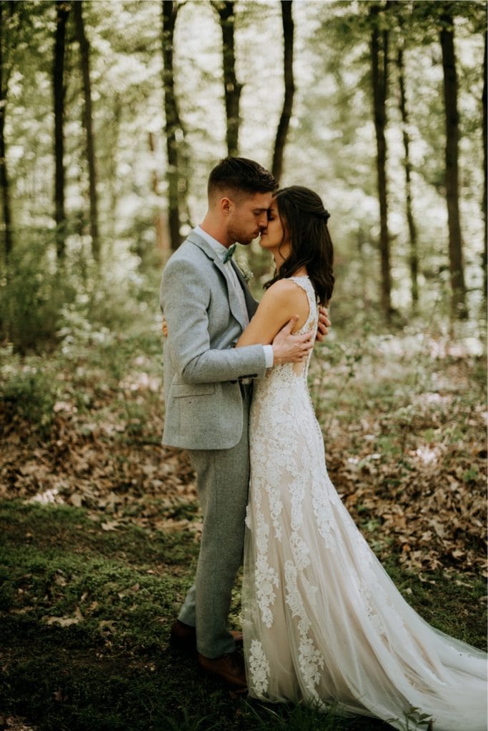 The sweetest first look in the forest before a wedding ceremony at the Wilds Venue in Bloomington, Indiana. The couple is embracing as light dapples the forest floor around them.