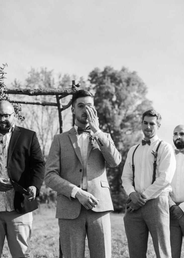 A groom crying as he sees his bride walking down the aisle, he's standing under their handmade wooden ceremony arch decorated with florals, during their outdoor wedding ceremony in the summer at The Wilds Venue in Bloomington, Indiana.