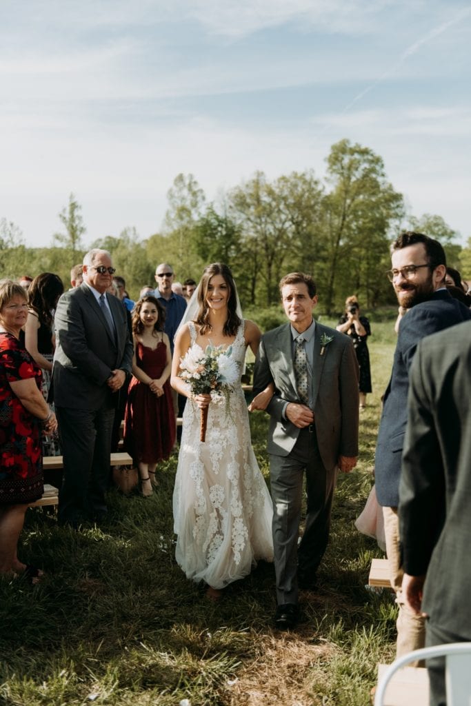 Bride walking down the aisle with her father during a summer outdoor wedding ceremony at the Wilds Venue.