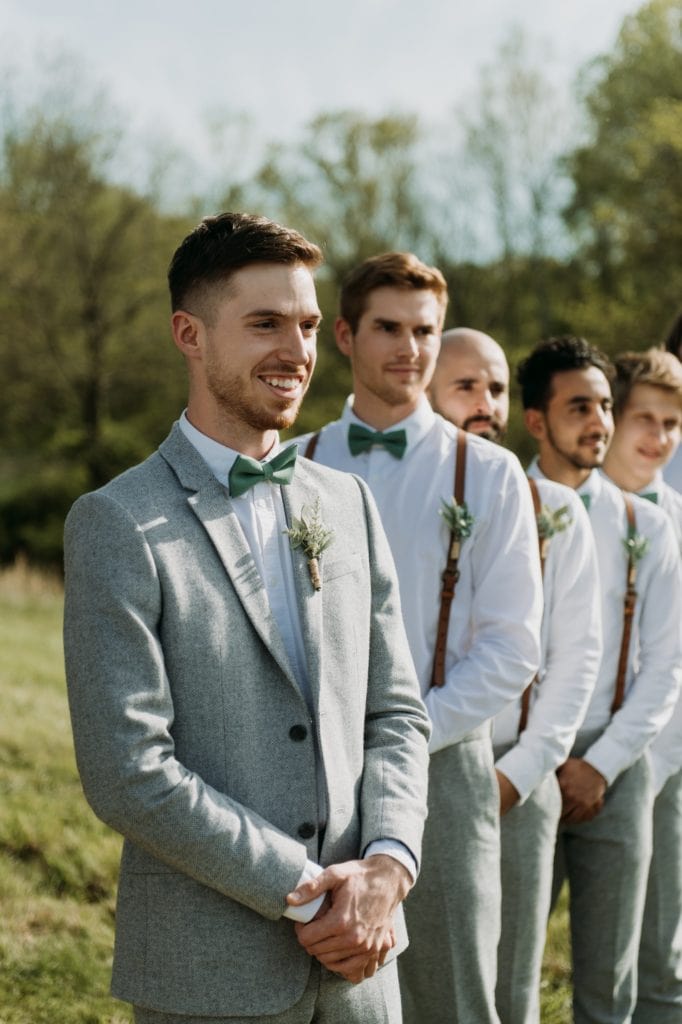 A groom smiling as he sees his bride walking down the aisle, he's standing under their handmade wooden ceremony arch decorated with florals, during their outdoor wedding ceremony in the summer at The Wilds Venue in Bloomington, Indiana.