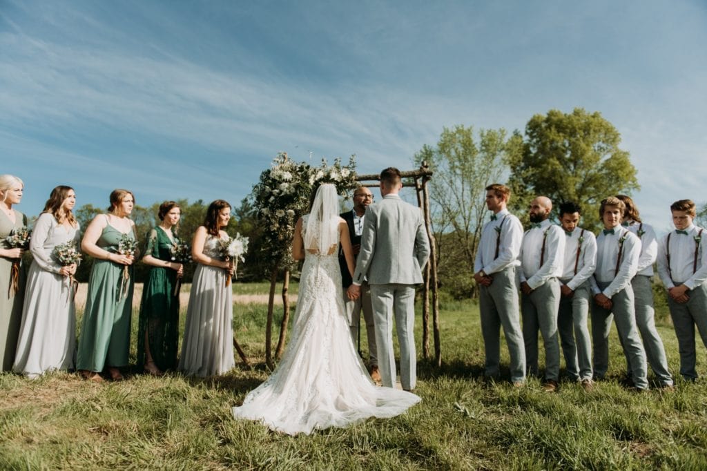 Bride and groom holding hands during the ceremony, they're standing under their handmade wooden ceremony arch decorated with florals, during their outdoor wedding ceremony in the summer at The Wilds Venue in Bloomington, Indiana.