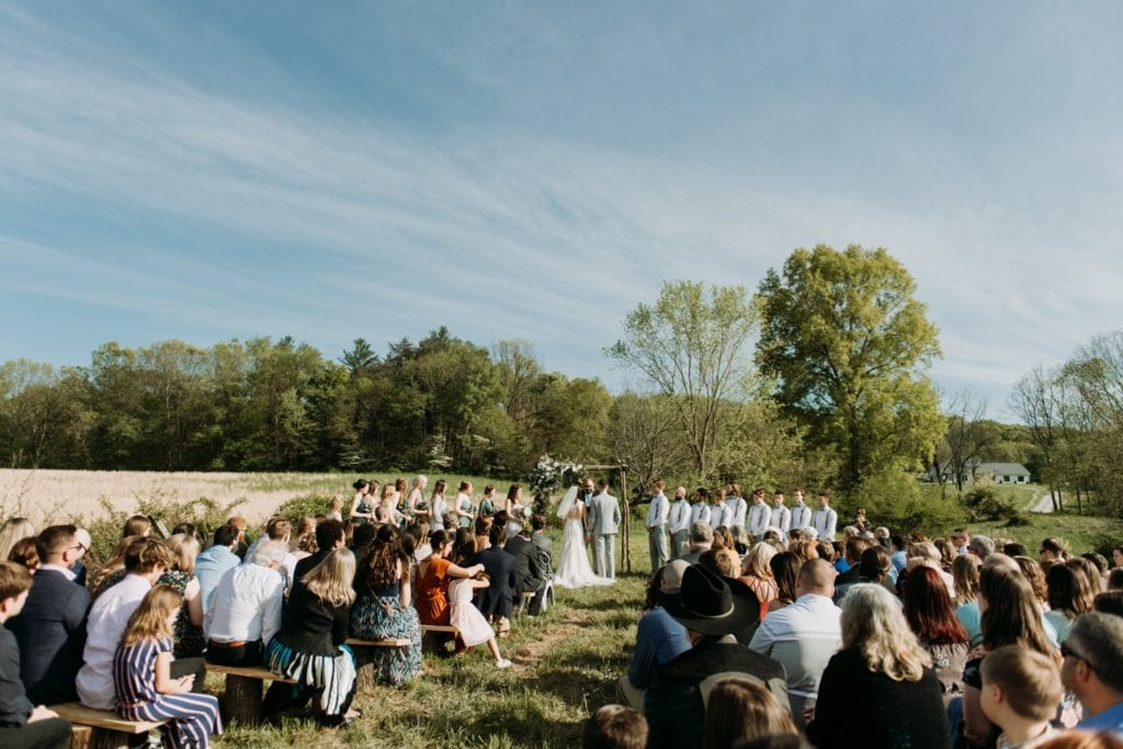 Wide angle shot of bride and groom holding hands during the ceremony, they're standing under their handmade wooden ceremony arch decorated with florals, during their outdoor wedding ceremony in the summer at The Wilds Venue in Bloomington, Indiana. Lush trees and a field and a lake are in the background, a perfect setting for an intimate summer wedding.