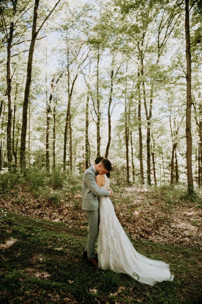 The sweetest first look in the forest before a wedding ceremony at the Wilds Venue in Bloomington, Indiana. The bride and groom are embracing each other as light dapples the forest floor around them.