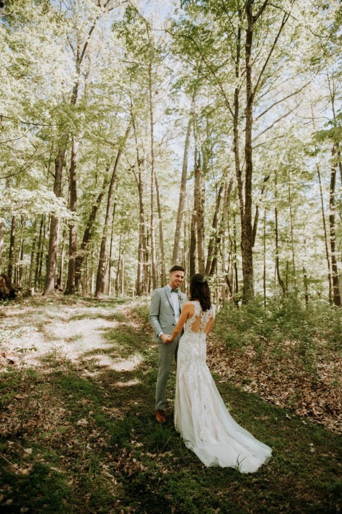 The sweetest first look in the forest before a wedding ceremony at the Wilds Venue in Bloomington, Indiana. The bride and groom are holding and hands and looking at each other as light dapples the forest floor around them.