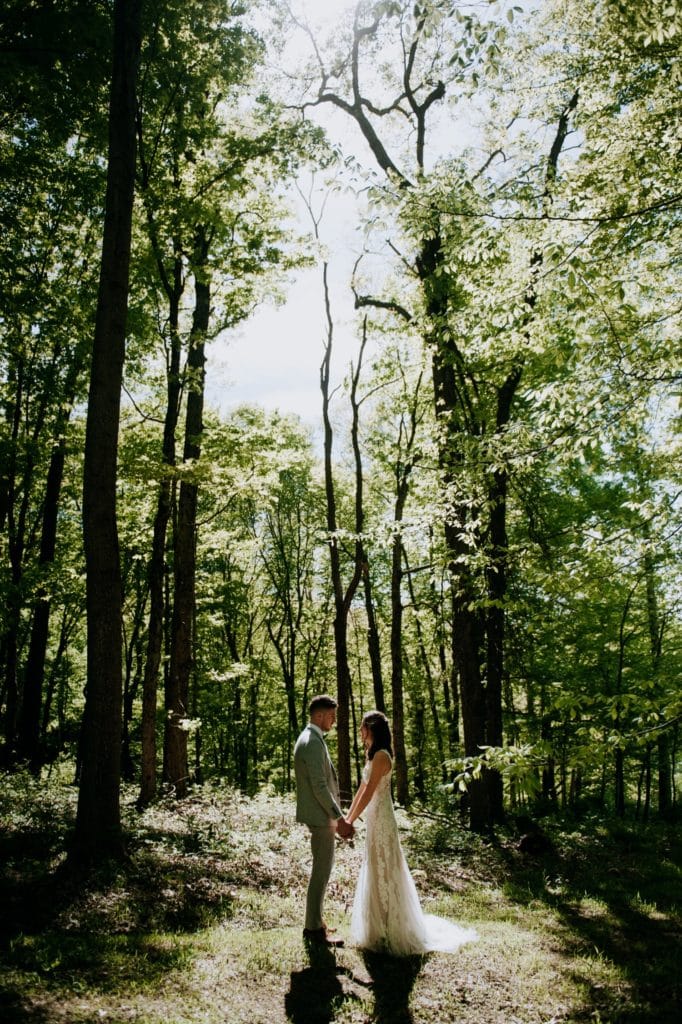 The sweetest first look in the forest before a wedding ceremony at the Wilds Venue in Bloomington, Indiana. The bride and groom are holding hands and looking at each other, standing in the light coming through the trees.