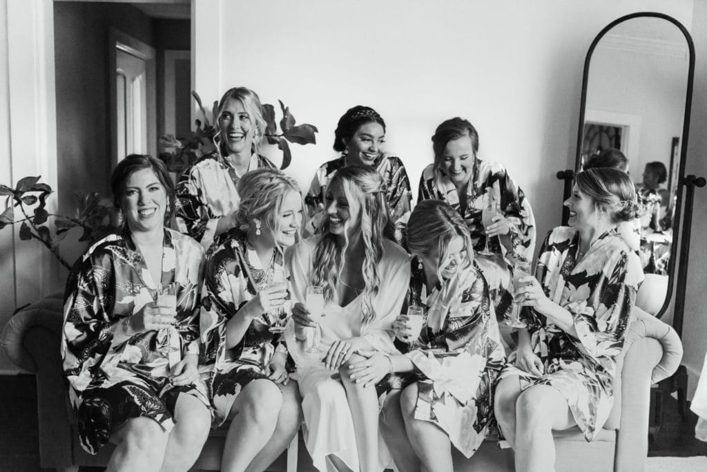A bride and her bridesmaids in their matching robes, clinking their champagne glasses, and sitting on a couch in the bridal suite at Mustard Seed Gardens in Indiana
