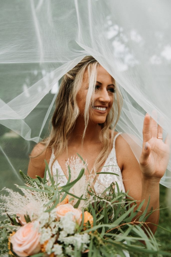 Bridal portraits under the veil. The bride is in the middle of the frame, holding her bouquet and smiling before the boho wedding ceremony at Mustard Seed Gardens.