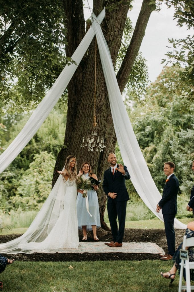 Bride and groom standing underneath a large oak tree during their boho summer wedding ceremony at Mustard Seed Gardens.