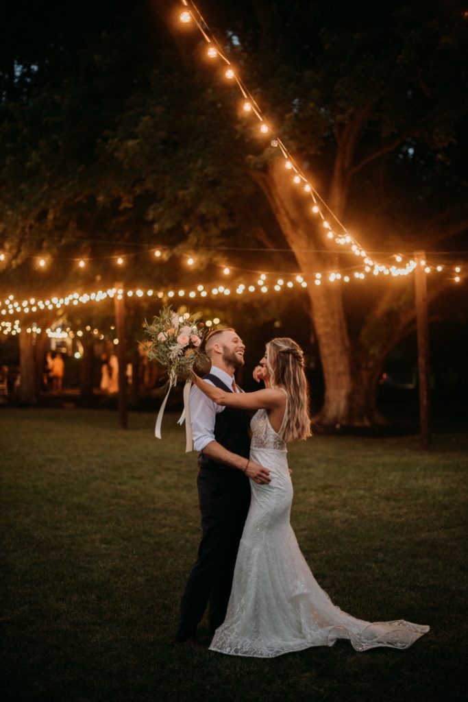 Bride and groom dancing and laughing underneath string lights at Mustard Seed Gardens