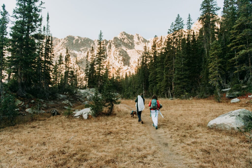 A couple hikes in their wedding attire after their elopement ceremony. Alpine lake hiking elopement ideas with their dog.