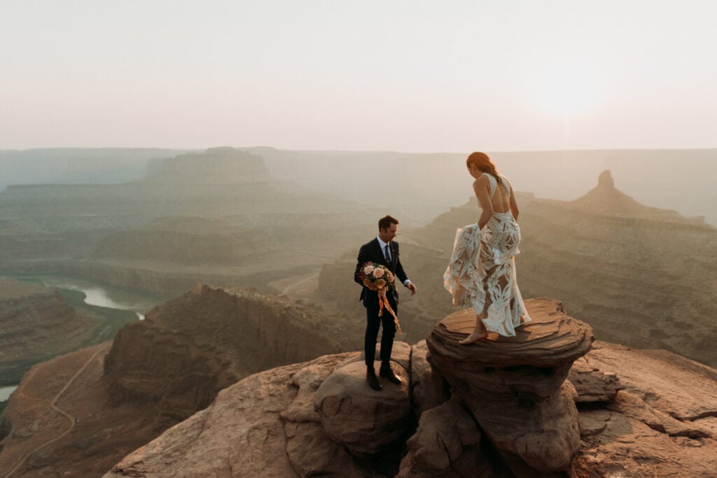 Bride and groom climbing on rocks in front of a canyon in moab utah. moab elopement photographer, how to elope in moab