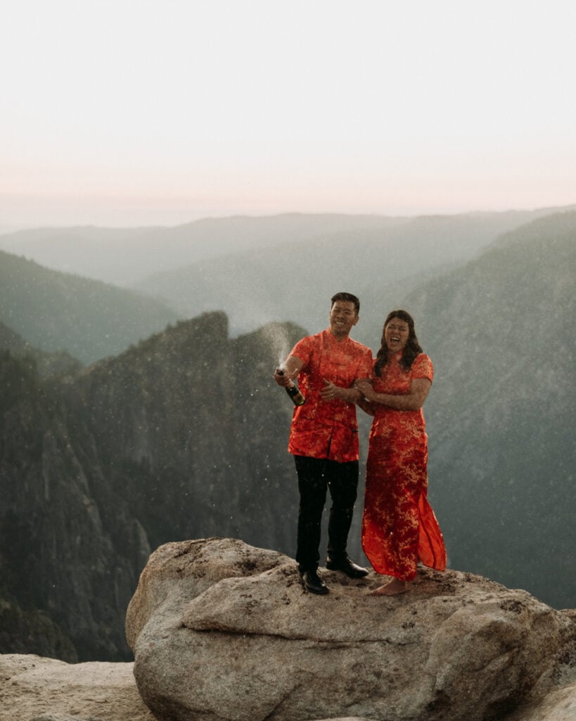 A couple wearing traditional Chinese wedding attire pops a bottle of champagne in front of a colorful sunset at Taft Point during their Yosemite elopement
