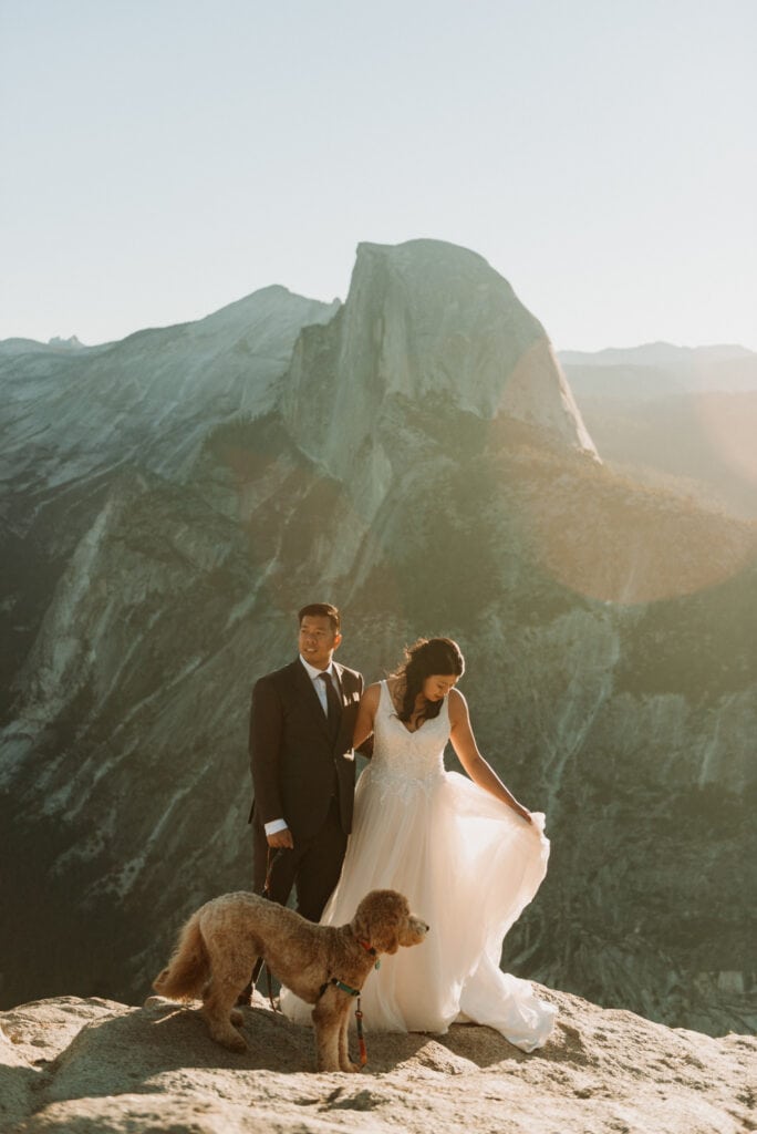 A wedding couple with their dog admires the landscape around them. Half dome is shining behind them right after sunrise in Yosemite National Park on their elopement day