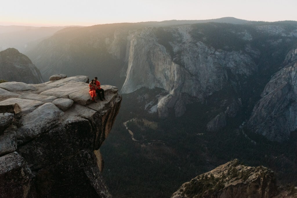 A couple in traditional Chinese wedding attire snuggles at Taft Point as the sun sets in the background in Yosemite National Park during their elopement