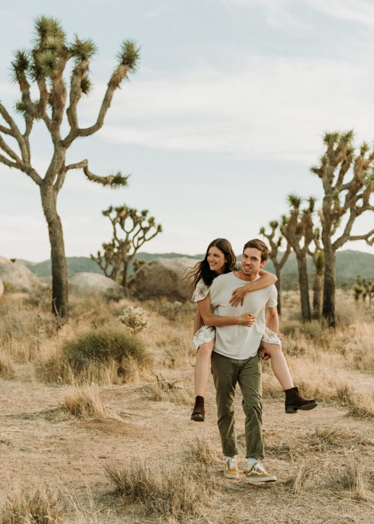 A woman getting a piggy back ride from her husband during their anniversary session in Joshua Tree california. Joshua tree national park engagement photos at cap rock