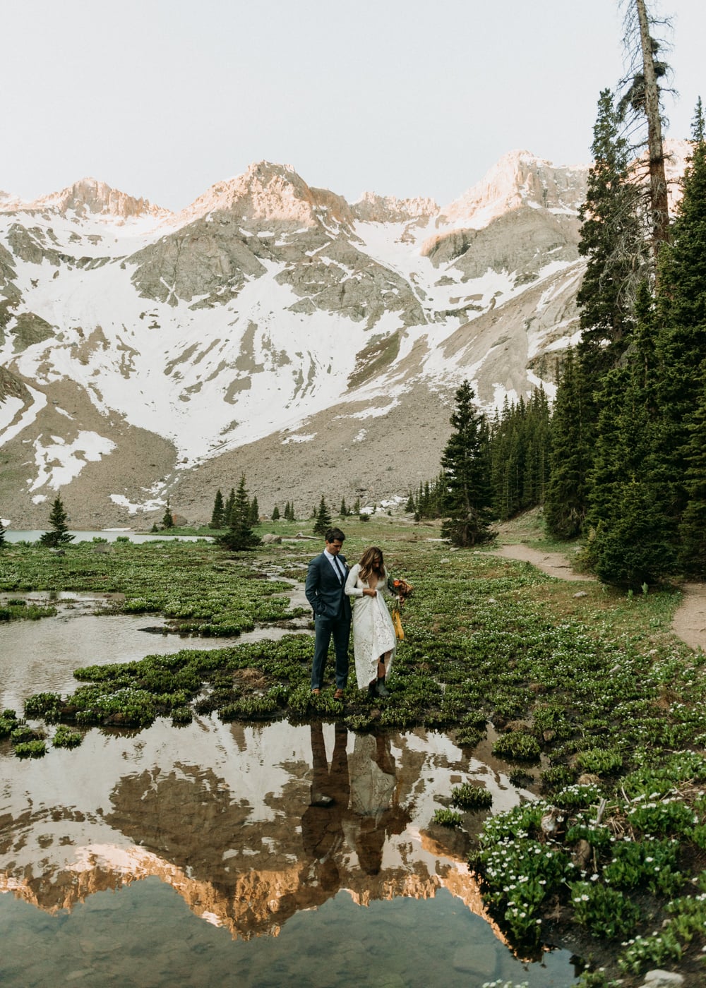A couple walking through wildflowers in front of an alpine lake and mountains in Telluride, Colorado on their elopement day.