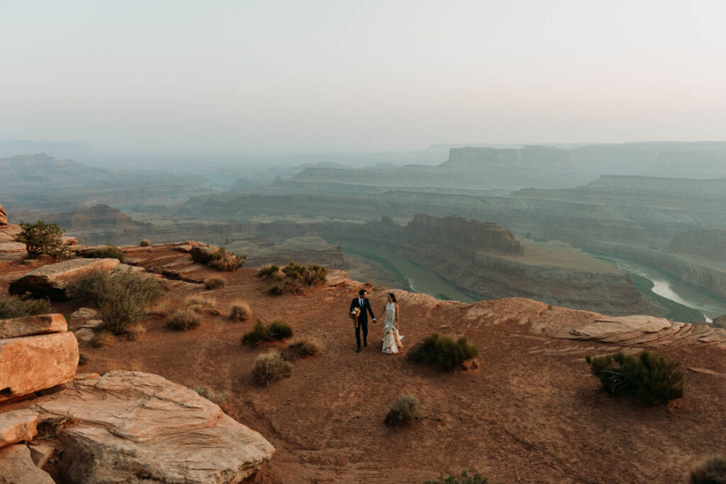 Couple walking along the edge of a desert canyon overlooking the Colorado River in Moab Utah at their micro wedding venue - Dead Horse Point State Park