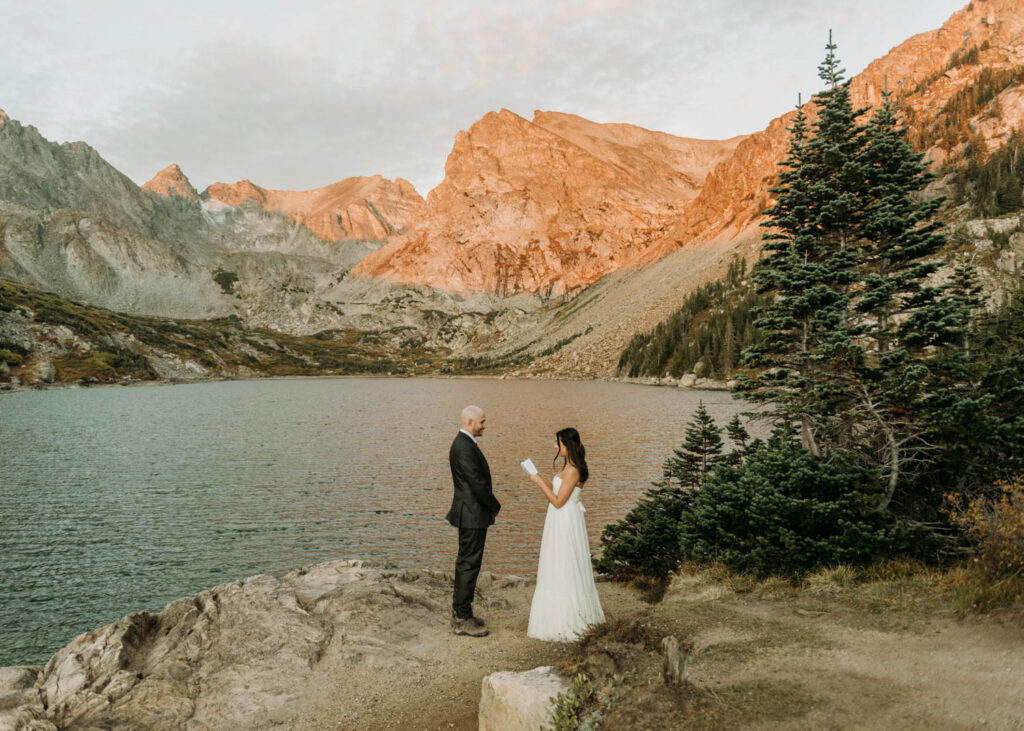 A couple stands in front of Lake Isabelle and the jagged mountain peaks as the alpenglow colors the mountains orange during their elopement