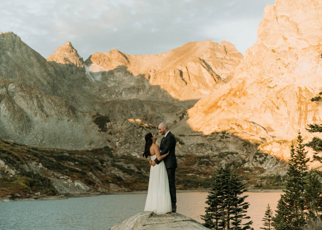 a couple in wedding attire stands on a huge rock at lake isabelle in colorado
