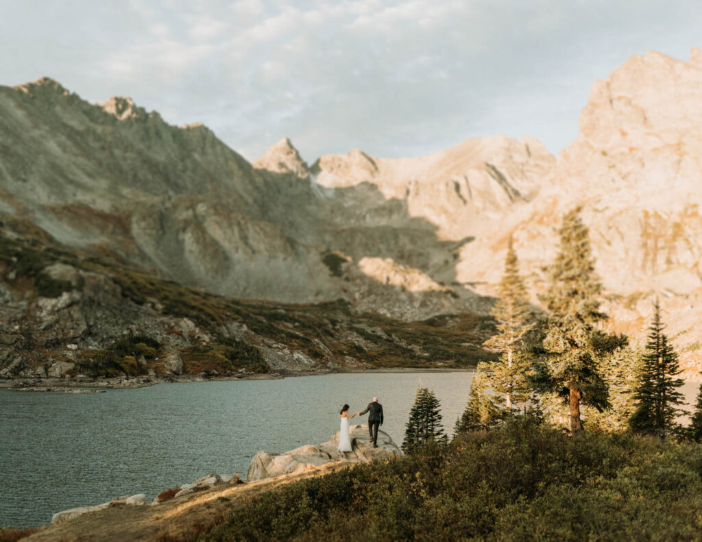 A couple holds hands walking along a huge rock at lake Isabelle during their hiking elopement