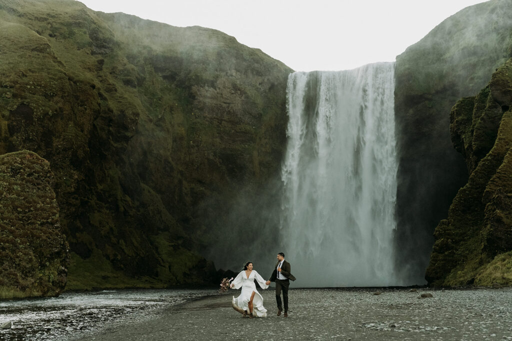 A couple running under the iconic Skogafoss waterfall, one of the best places to elope in Iceland, during their elopement.