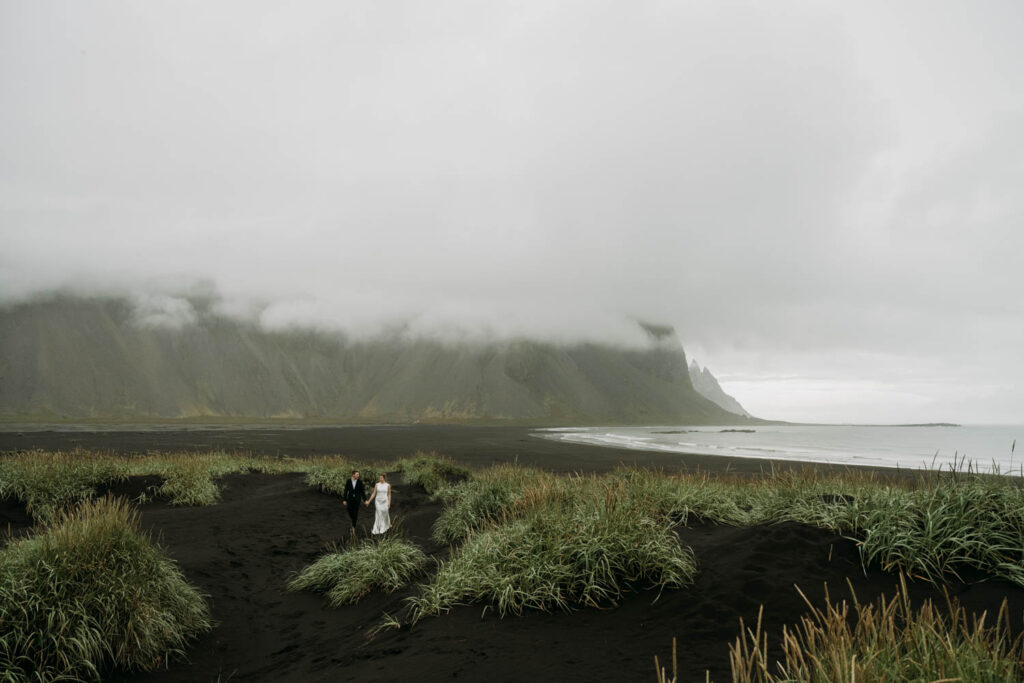 Walking through the black sand beach, a couple is hand in hand before their ceremony for their Stokksnes elopement